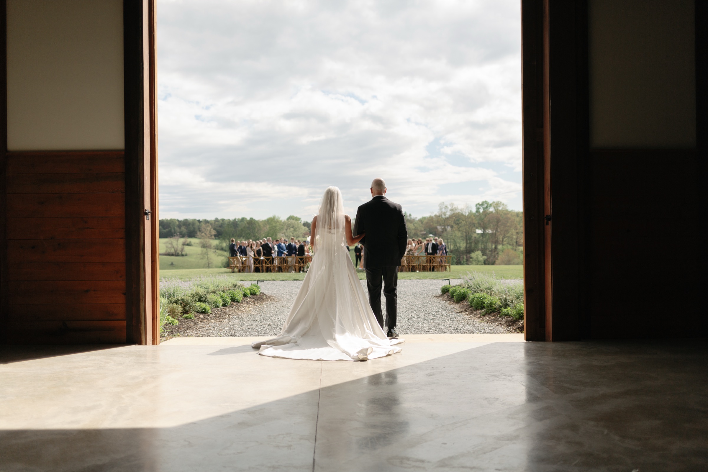 The bride and her father step out through the open barn doors toward the ceremony on the lawn