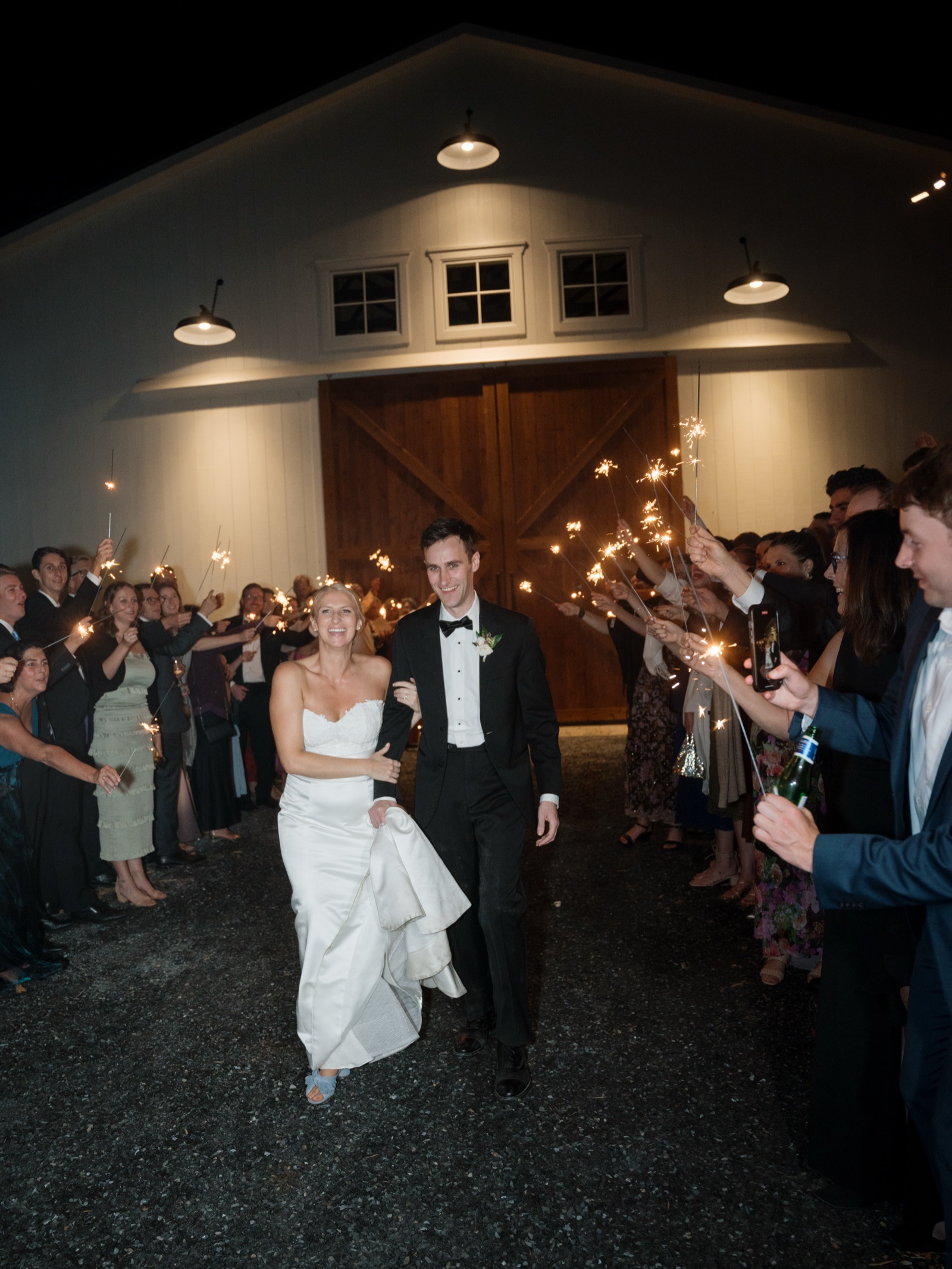 A couple walking out between two lines of guests holding sparklers, the barn doors lit behind them